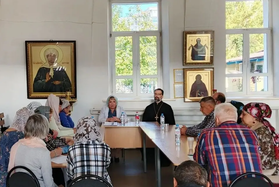 Священник и специалист сидят во главе стола, вокруг — участники круглого стола о помощи зависимым, на стене иконы
