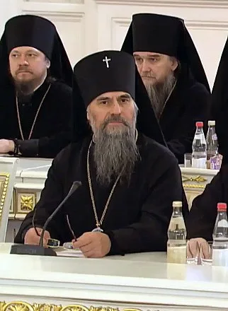 Архиерей в чёрном клобуке и панагии за столом среди других священнослужителей